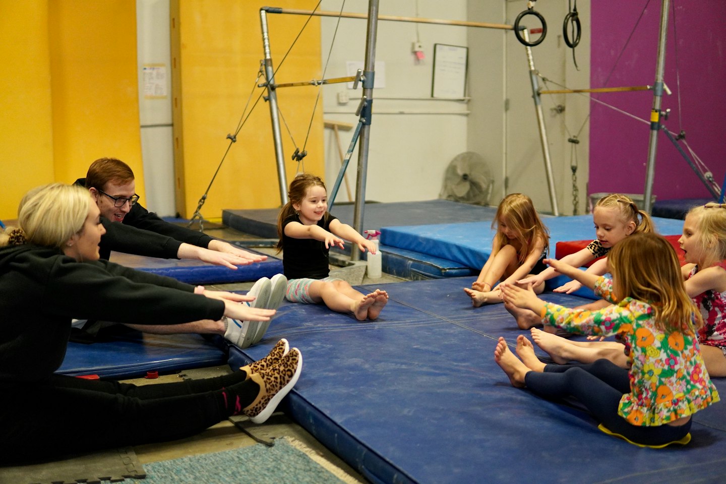 Deti a dospelí sa naťahujú na modrých podložkách v gymnastickej hale.